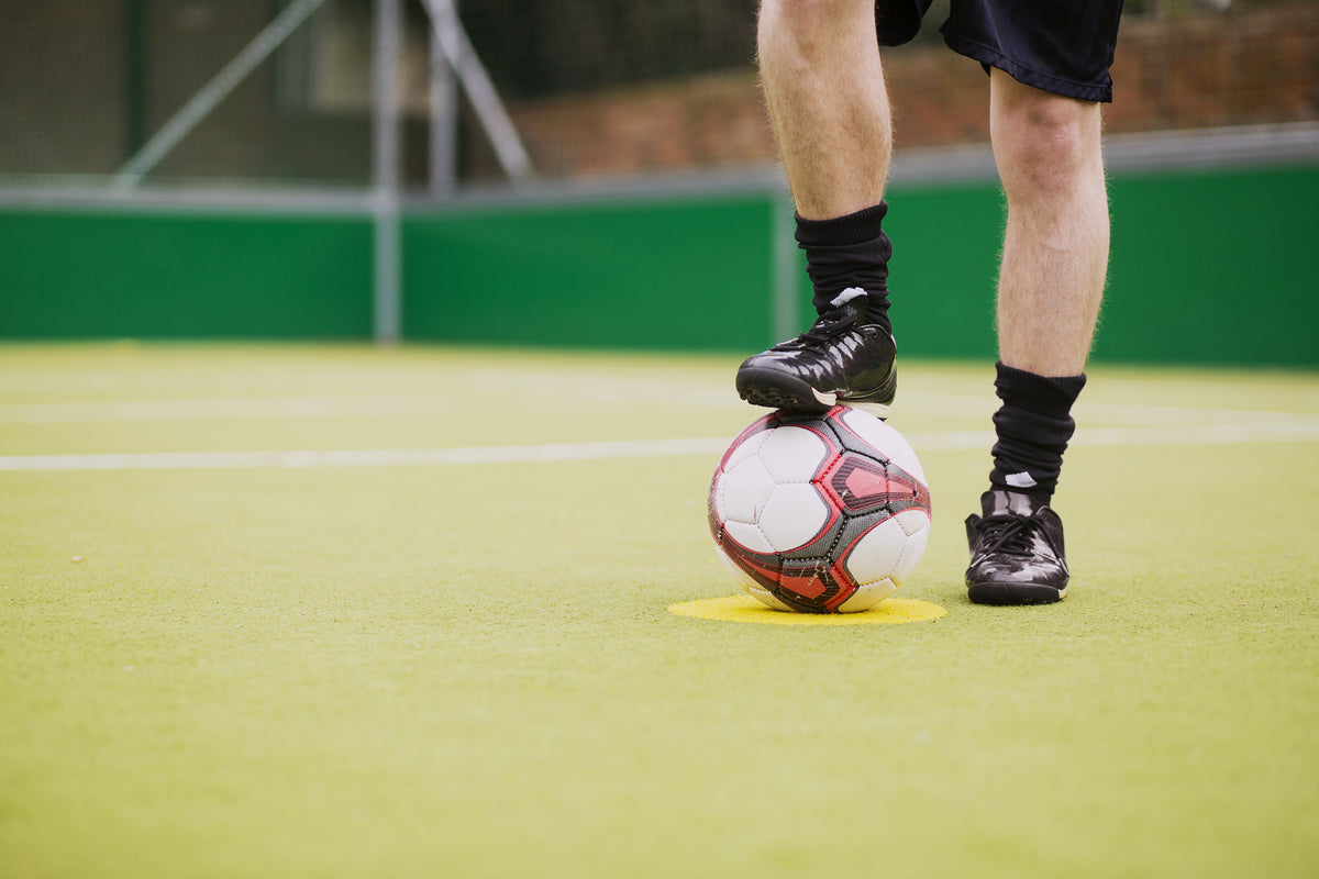 A close-up action shot of a soccer player's legs and feet on a green turf field, balancing a white soccer ball with red and black patterns on their foot.