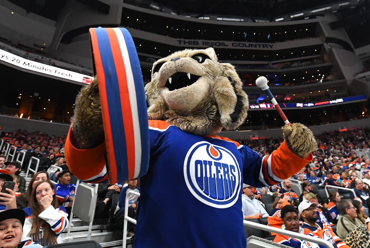 An Edmonton Oilers mascot in a blue and orange hockey jersey stands in an arena, holding a large blue, white, and orange round sign and a small stick with a white ball.